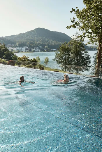 Paar schwimmt im Infinity-Pool mit Blick auf See und bewaldete Berge