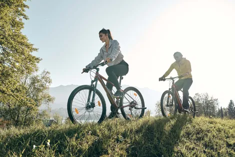 Couple riding mountain bikes on grassy trail in sunlight with forest and mountains in background