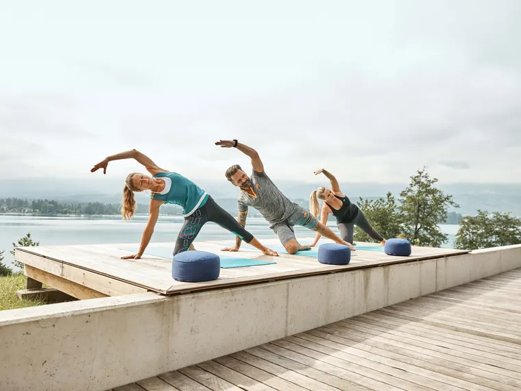 Drei Personen machen Yoga in der Seitstützposition auf einem Holzdeck am See