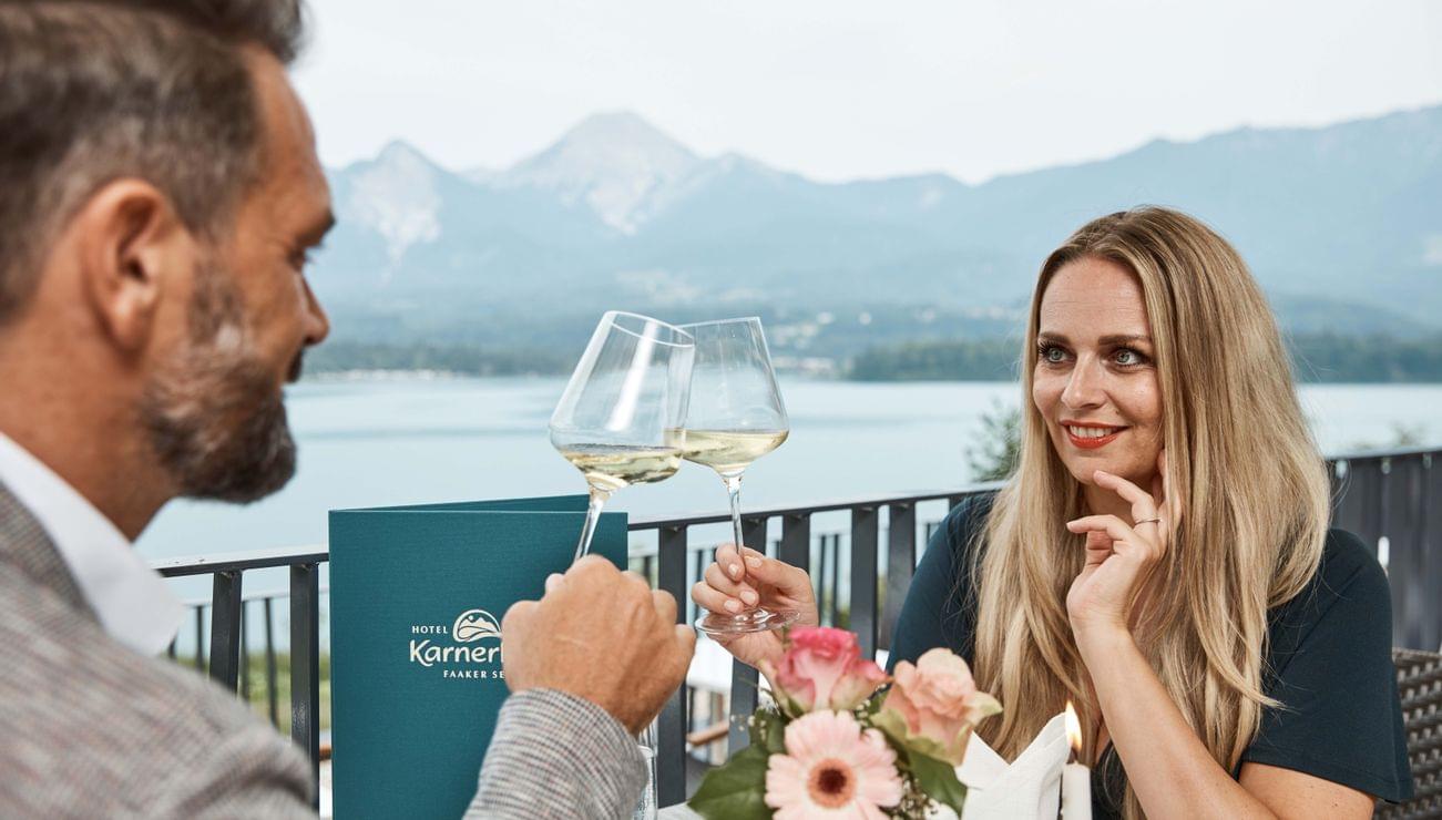 Paar stößt mit Weißwein auf Hoteldachterrasse mit Blick auf See und Berge an