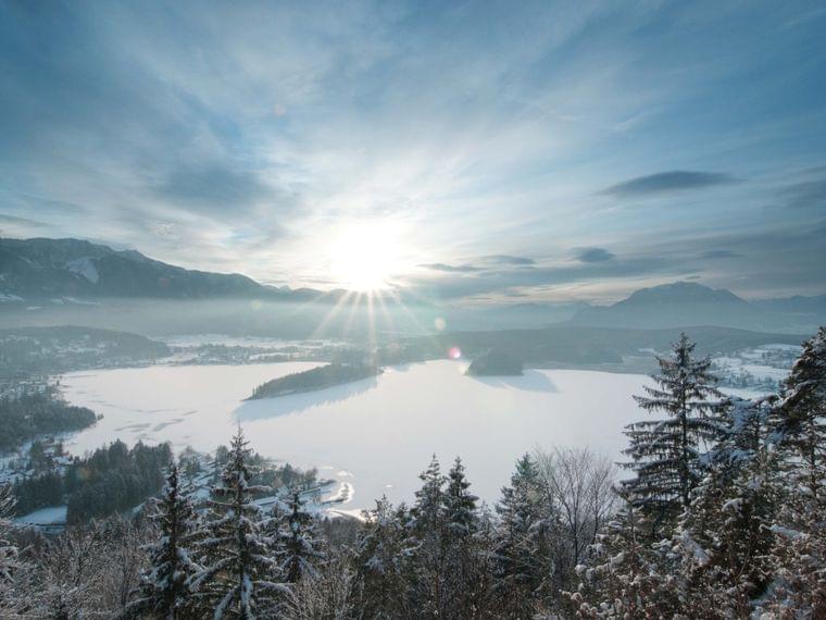 Snow-covered lake surrounded by pine trees and mountains at winter sunrise