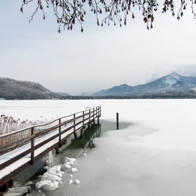 Verschneiter Holzsteg auf zugefrorenem Alpensee mit Blick auf verschneite Berge