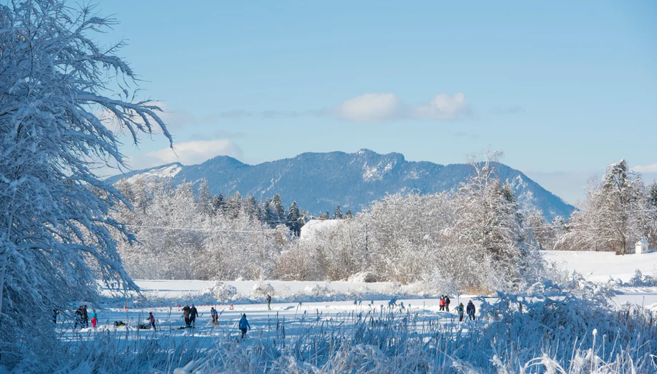 Menschen beim Eislaufen auf einem zugefrorenen See mit verschneiten Bäumen und Bergen