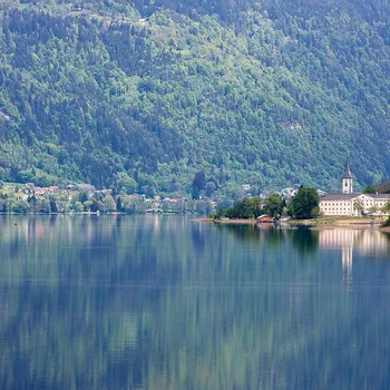 See mit Alpenort und Kirche, die sich im ruhigen Wasser vor bewaldeten Bergen spiegeln