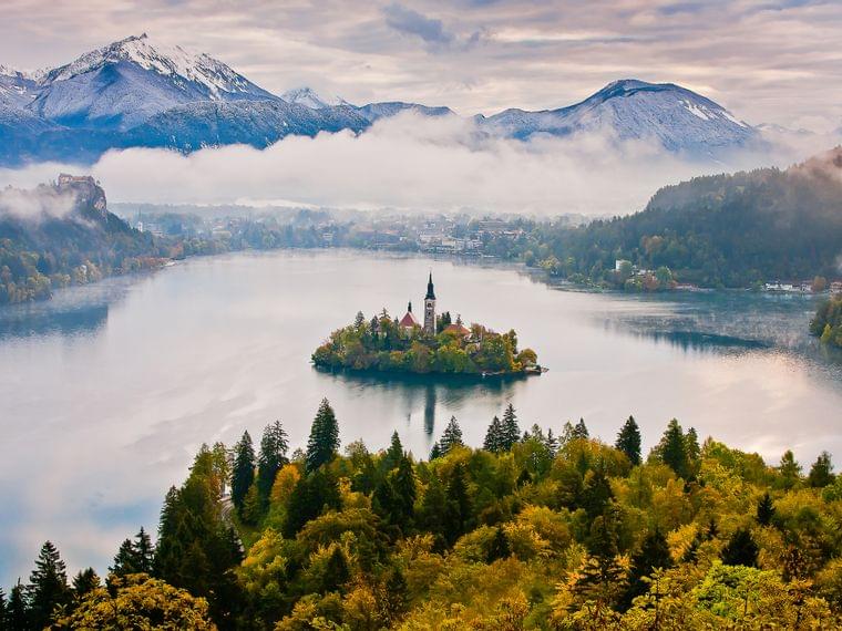 Lago di Bled con chiesa sull'isola, circondato da boschi autunnali e Alpi Giulie innevate