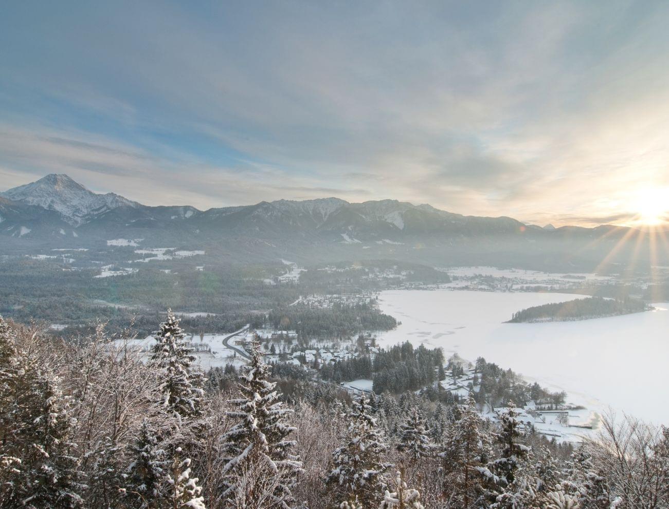 Verschneite Alpenlandschaft mit Wald, zugefrorenem See und aufgehender Sonne über Bergen